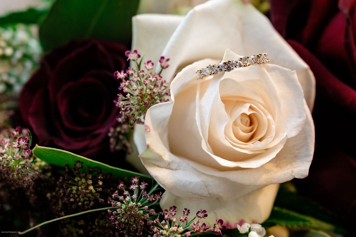 A series of delicate cream and white roses arranged in a row with dark burgundy accents and baby's breath flowers.