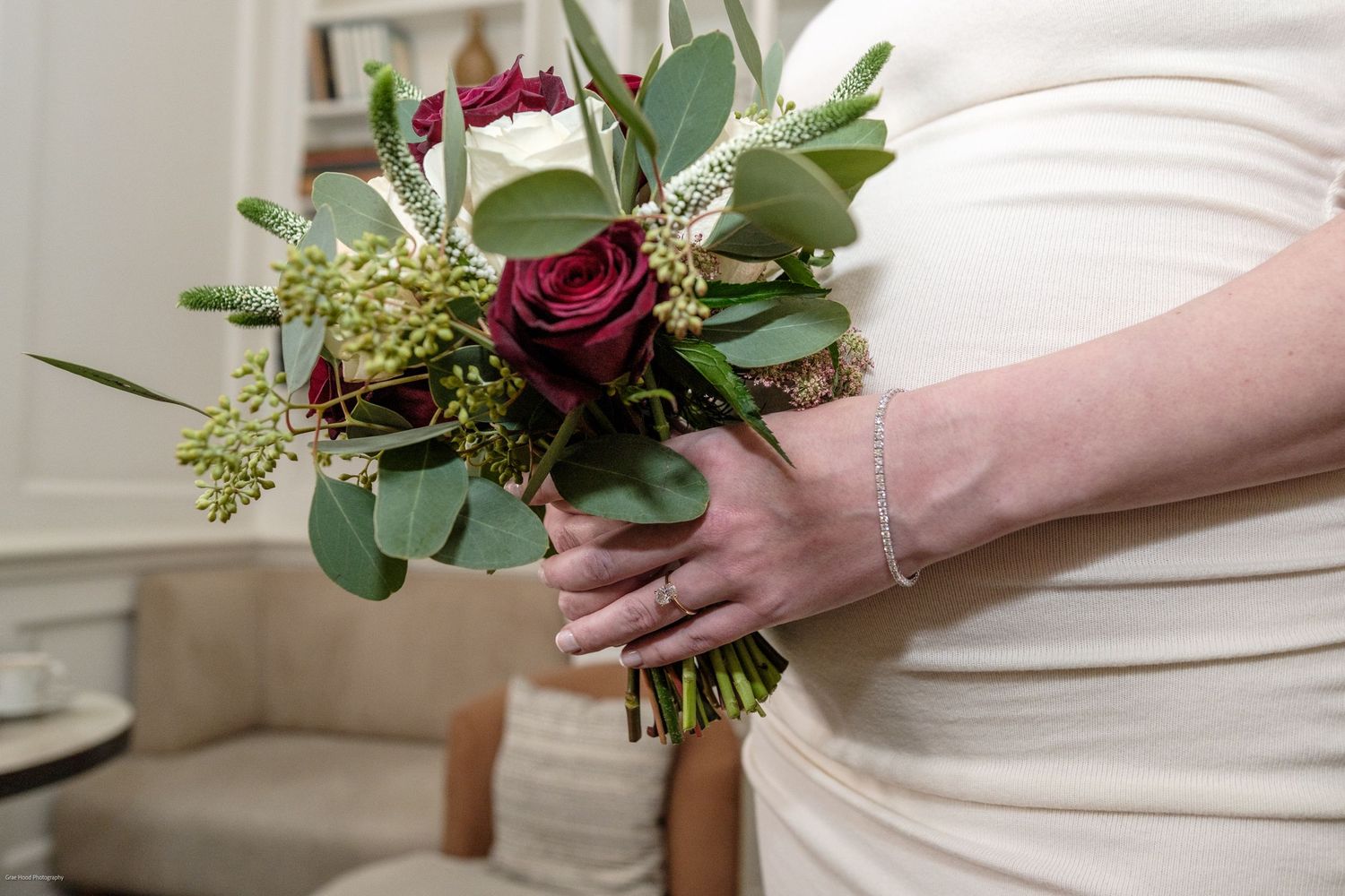 A hand holds a rustic wedding bouquet featuring deep red roses, greenery, and delicate white flowers against a white background.