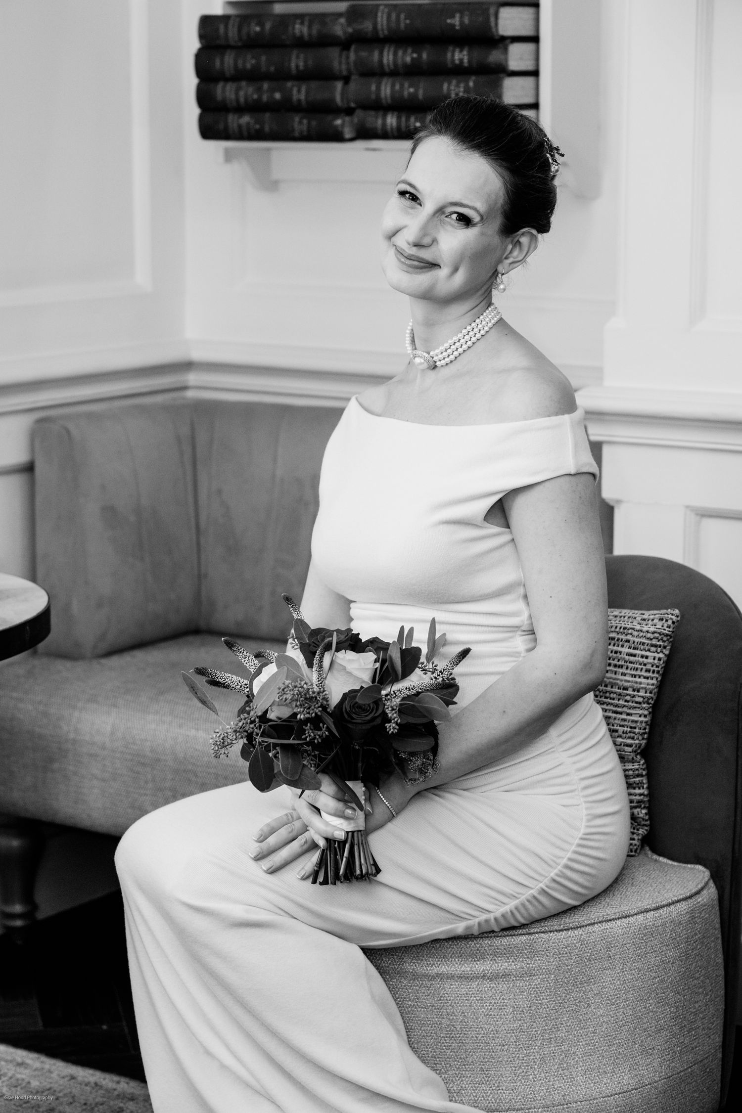 A black and white portrait of someone in an elegant white dress holding a bouquet while seated.