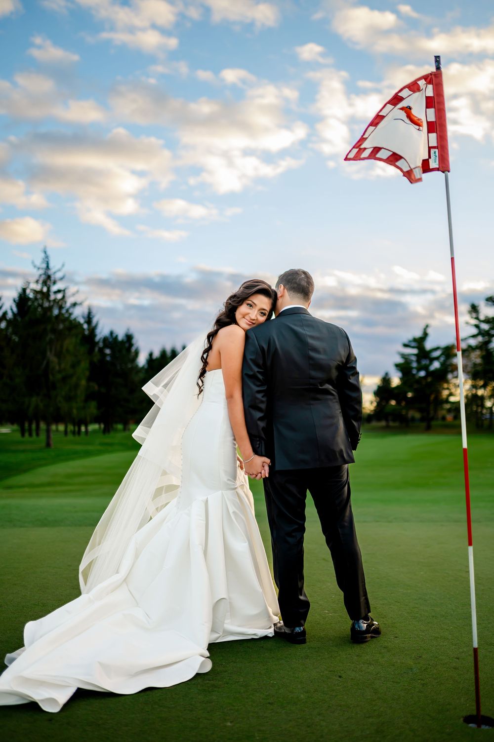 A couple in wedding attire playfully poses near a golf flag on a lush green course at sunset.