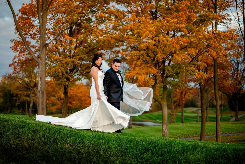 A couple in wedding attire walks through a scenic autumn park with vibrant orange and red foliage.