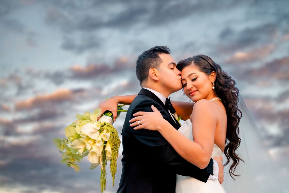 Couple embraces during sunset wedding photoshoot with dramatic cloudy sky and yellow bouquet.