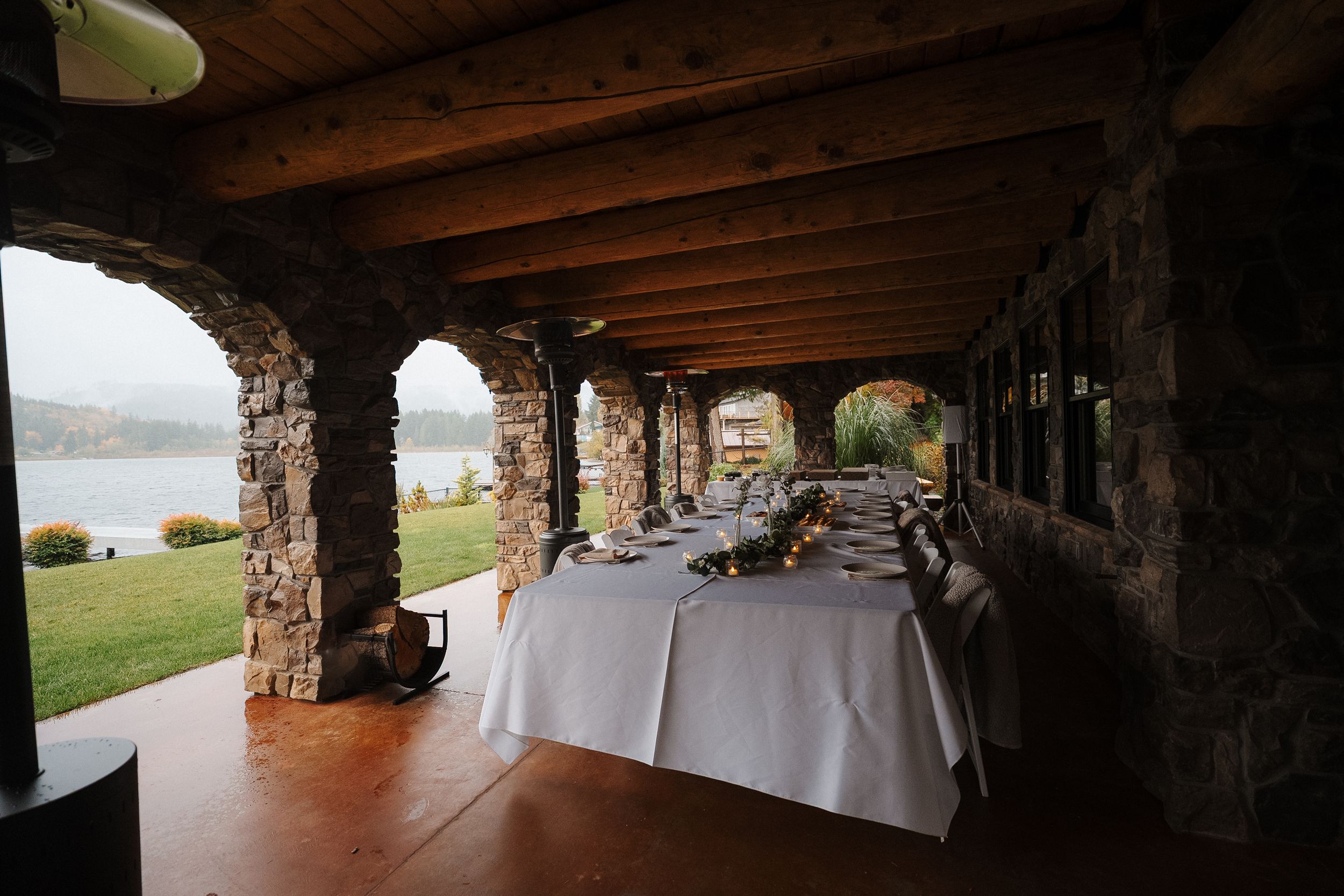 A rustic stone porch with wooden beams and white tablecloths set up for an outdoor dining event.