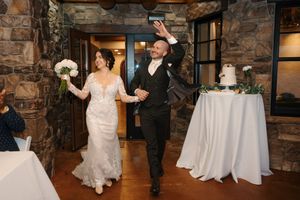 A couple makes their grand entrance at a rustic stone venue with a white wedding cake displayed on a nearby table.