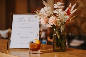 Wedding menu card displayed next to a craft cocktail and mason jar centerpiece with dried flowers on wooden table.