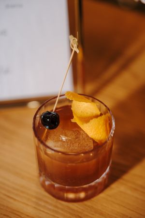 A craft cocktail garnished with an orange peel and dark cherry in a rocks glass on a wooden surface.