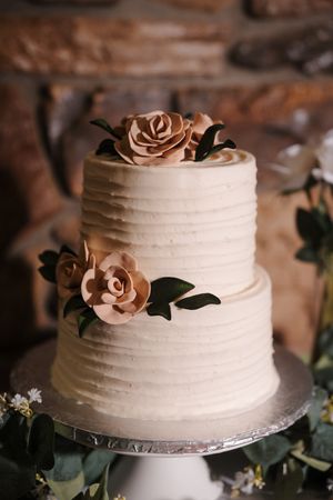 Elegant two-tier white wedding cake with blush pink sugar roses and green foliage on white cake stand.