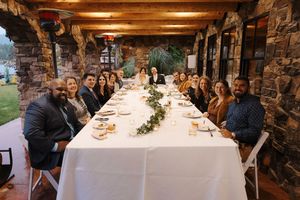 Long banquet table set for dinner inside rustic stone lodge with wooden beam ceiling and elegant place settings.