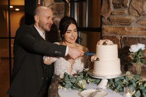 A couple cuts into their elegant two-tier white wedding cake decorated with greenery.