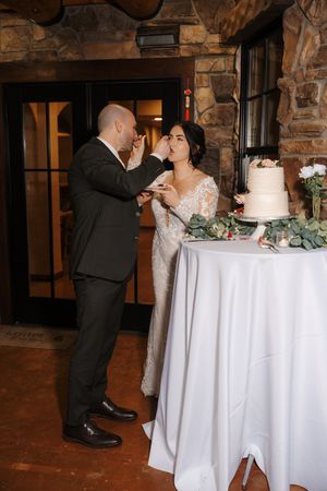 A couple in formal wedding attire shares a romantic moment while cutting their white tiered wedding cake at a rustic venue.