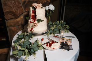 Elegant white wedding cake with burgundy floral accents and eucalyptus leaves displayed on white tablecloth.