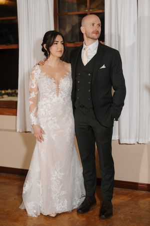 A couple in wedding attire stands together, with the bride wearing a white lace gown and the groom in a black suit.