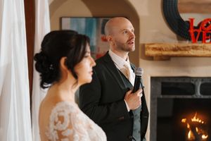A couple in formal wedding attire stands together in an elegant room with white drapes and warm lighting.