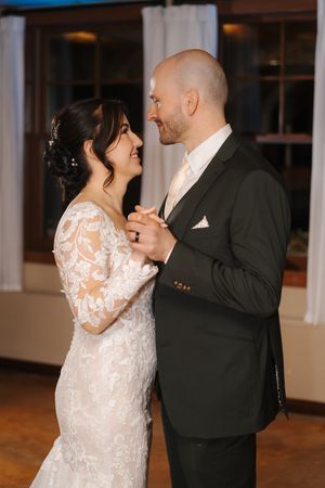 A couple shares their first dance at a wedding reception, with the bride wearing a white lace gown with long sleeves.