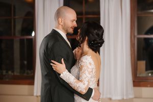 Newlyweds share their first dance in an elegant white reception hall with large windows.