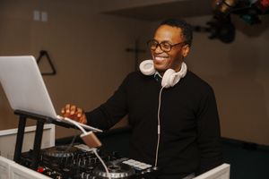 DJ wearing headphones and smiling while performing at a mixing console in a dimly lit venue.