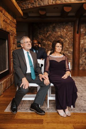 Couple sitting on rustic wooden bench against stone wall in formal attire at indoor venue.