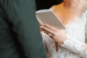 Bride in lace wedding dress holds a book while standing in front of a window with natural light.