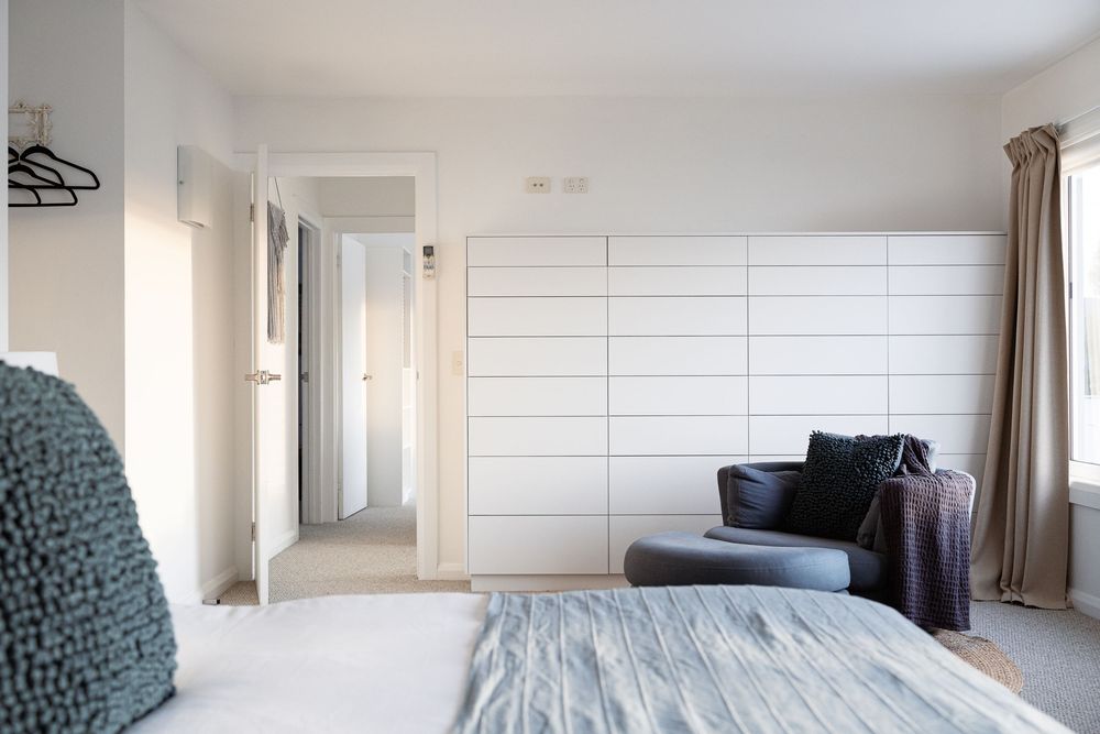 Modern minimalist bedroom with white walls, wooden floors, and large windows letting in natural light.
