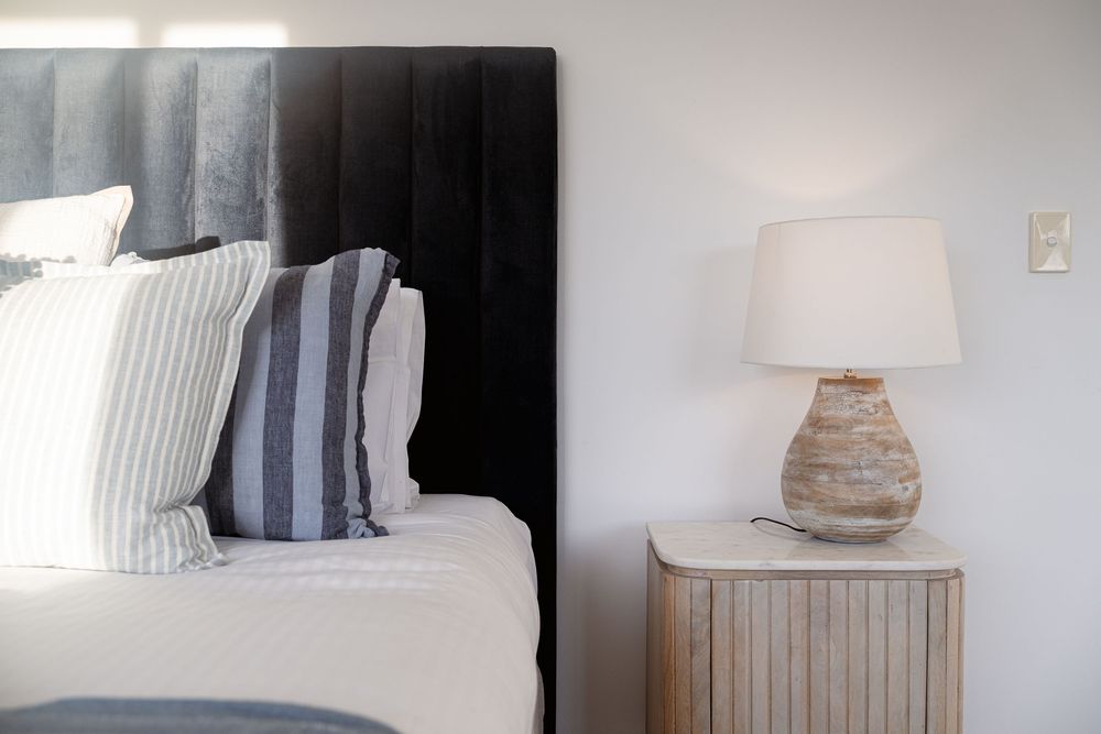 A cozy bedroom interior with striped pillows on a bed and a rustic table lamp on a wooden nightstand.
