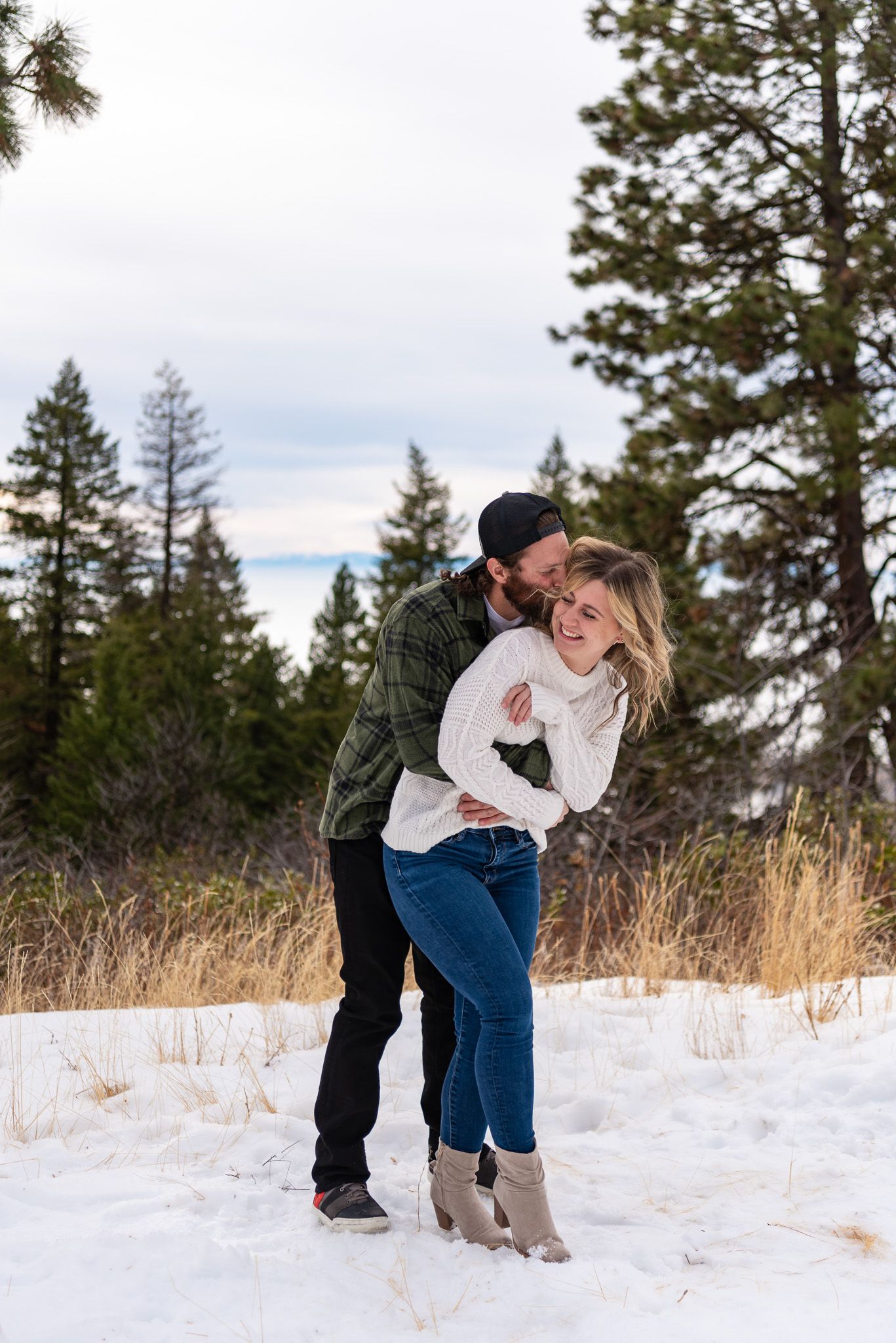 Loving couple embraces in snowy winter landscape with evergreen trees in background.
