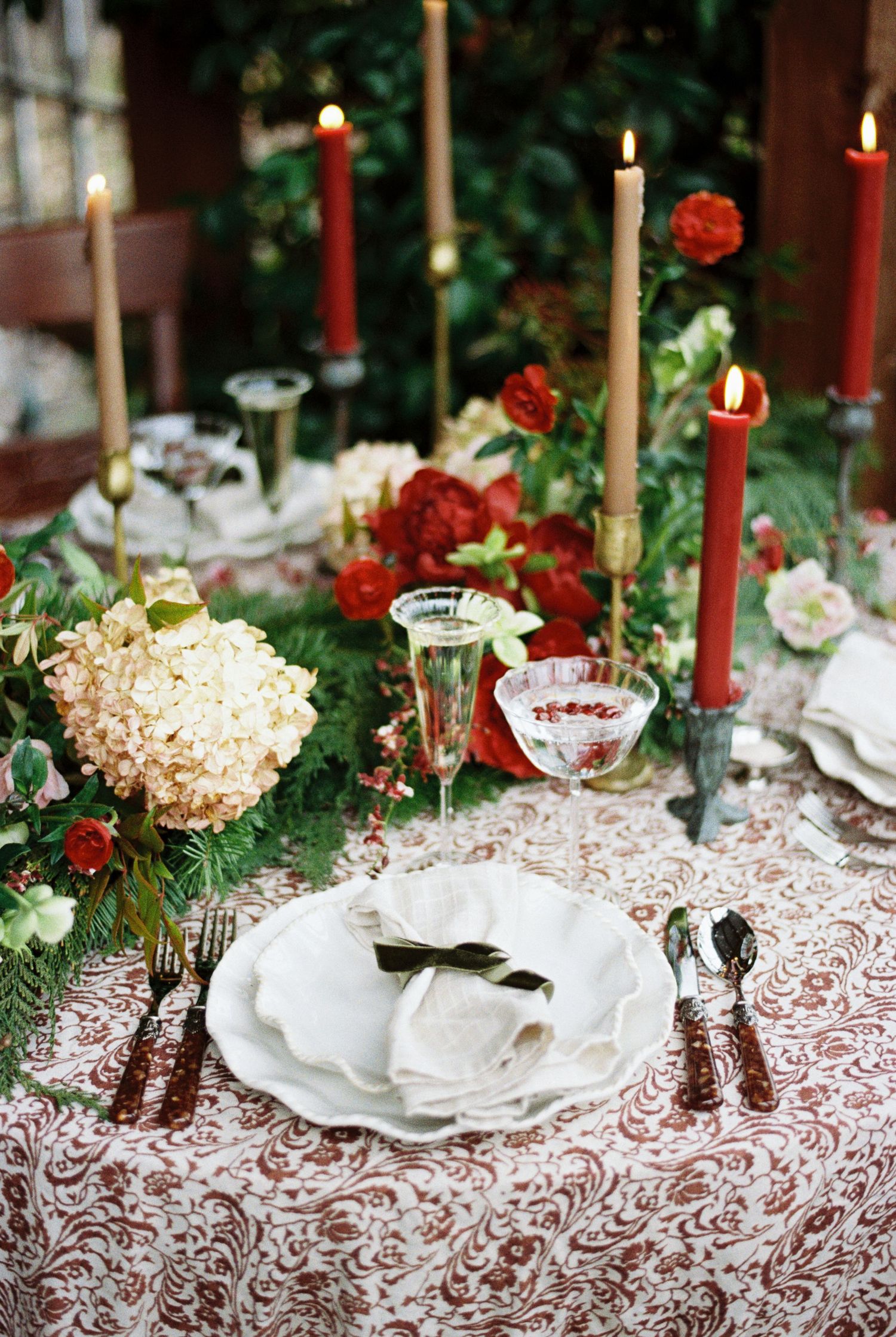 Elegant table setting with red candles, roses, and hydrangeas on lace tablecloth with vintage china and crystal glasses.