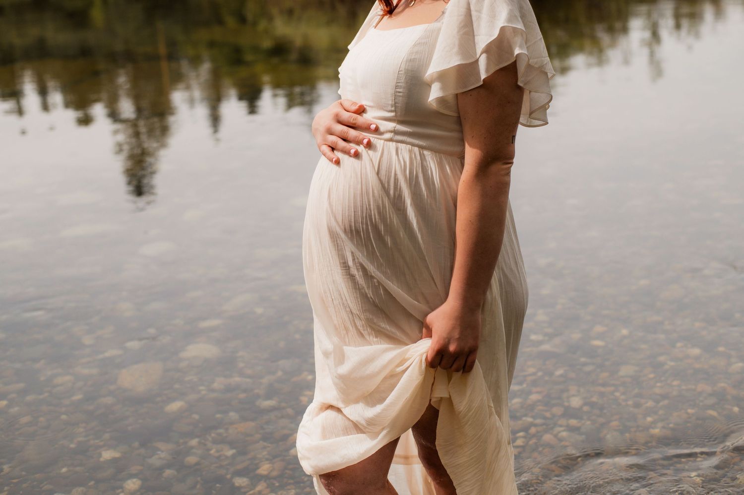 Pregnant woman in flowing white dress standing at edge of calm lake with reflections on water surface.