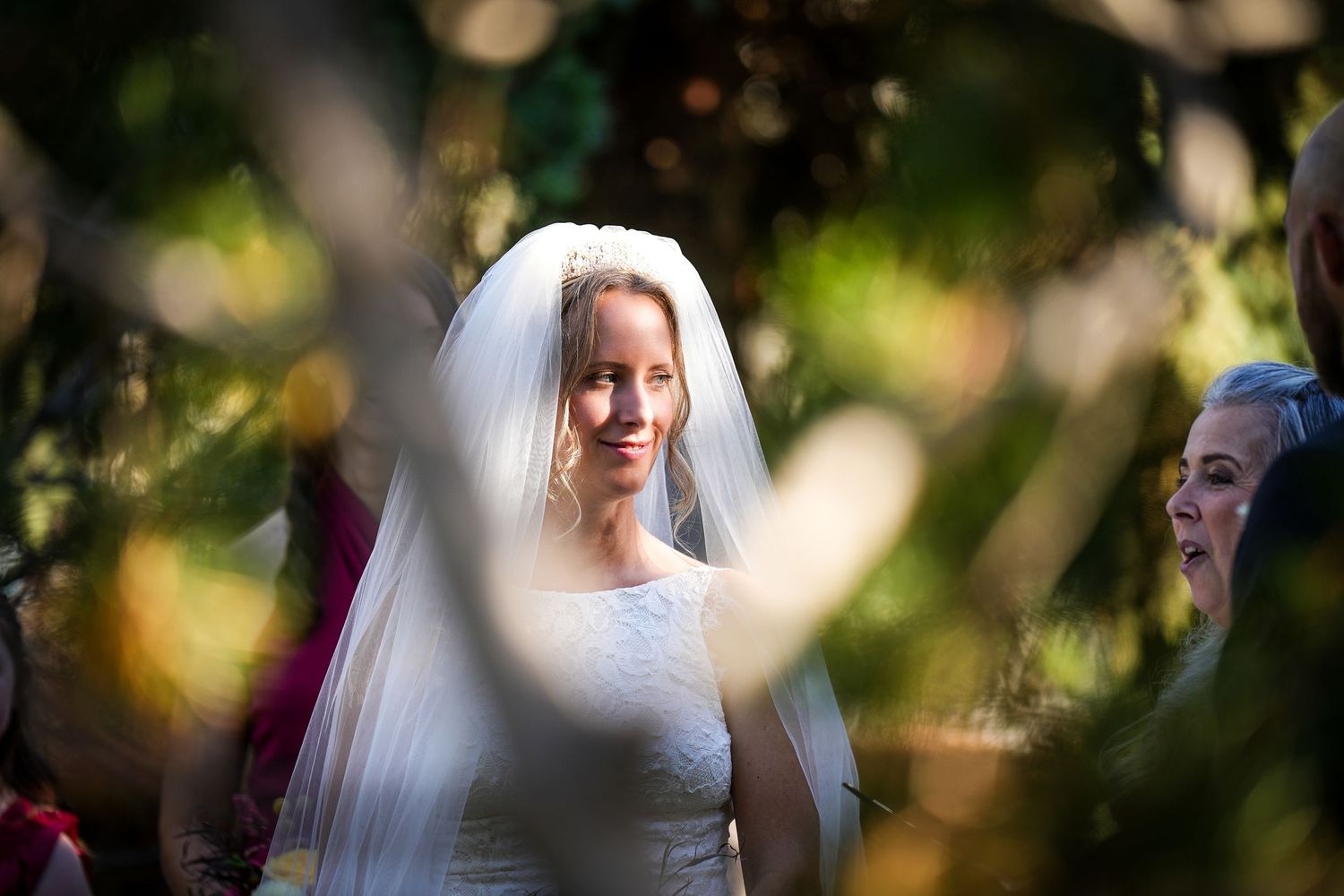 Artistic wedding photo through foliage capturing bride in white dress and veil.