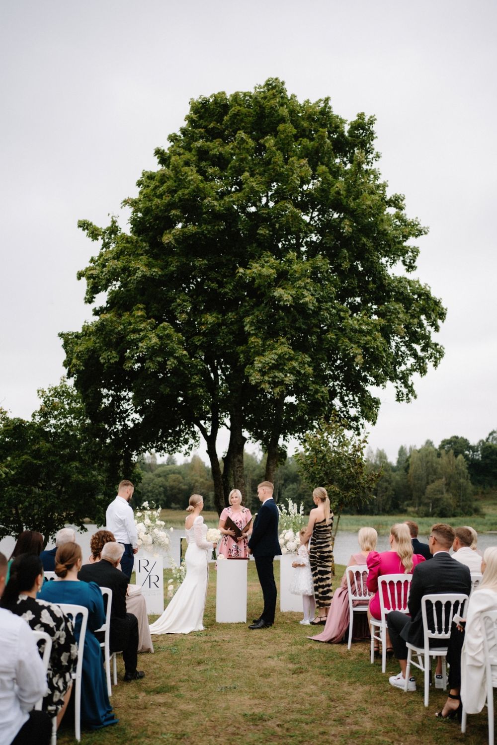 Wedding ceremony setup under large trees with white chairs and floral arrangements.