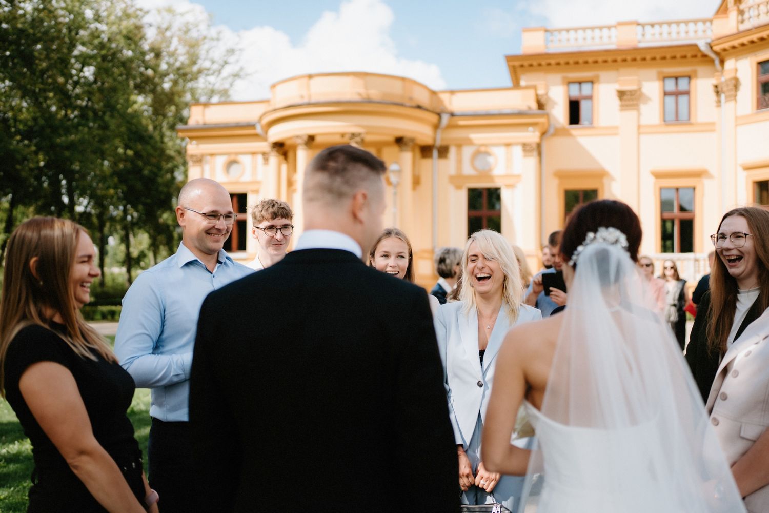 Outdoor wedding gathering in front of a grand yellow mansion with classical architecture.