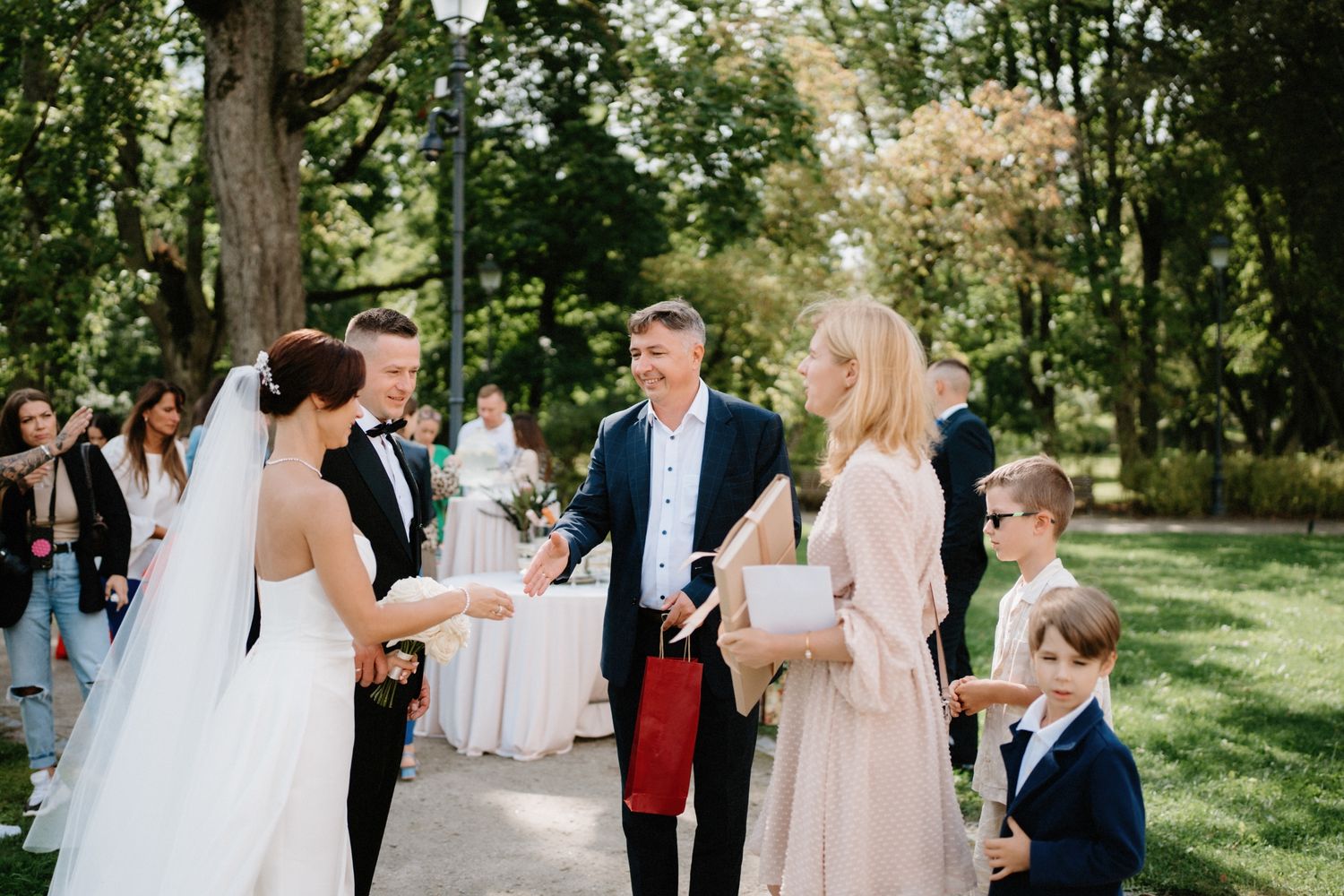 Wedding reception guests mingling on garden path surrounded by trees and sunlight.