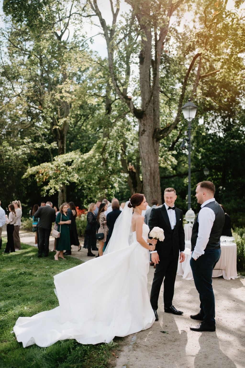 Stunning bride in flowing white gown at outdoor garden reception with golden sunlight filtering through trees.