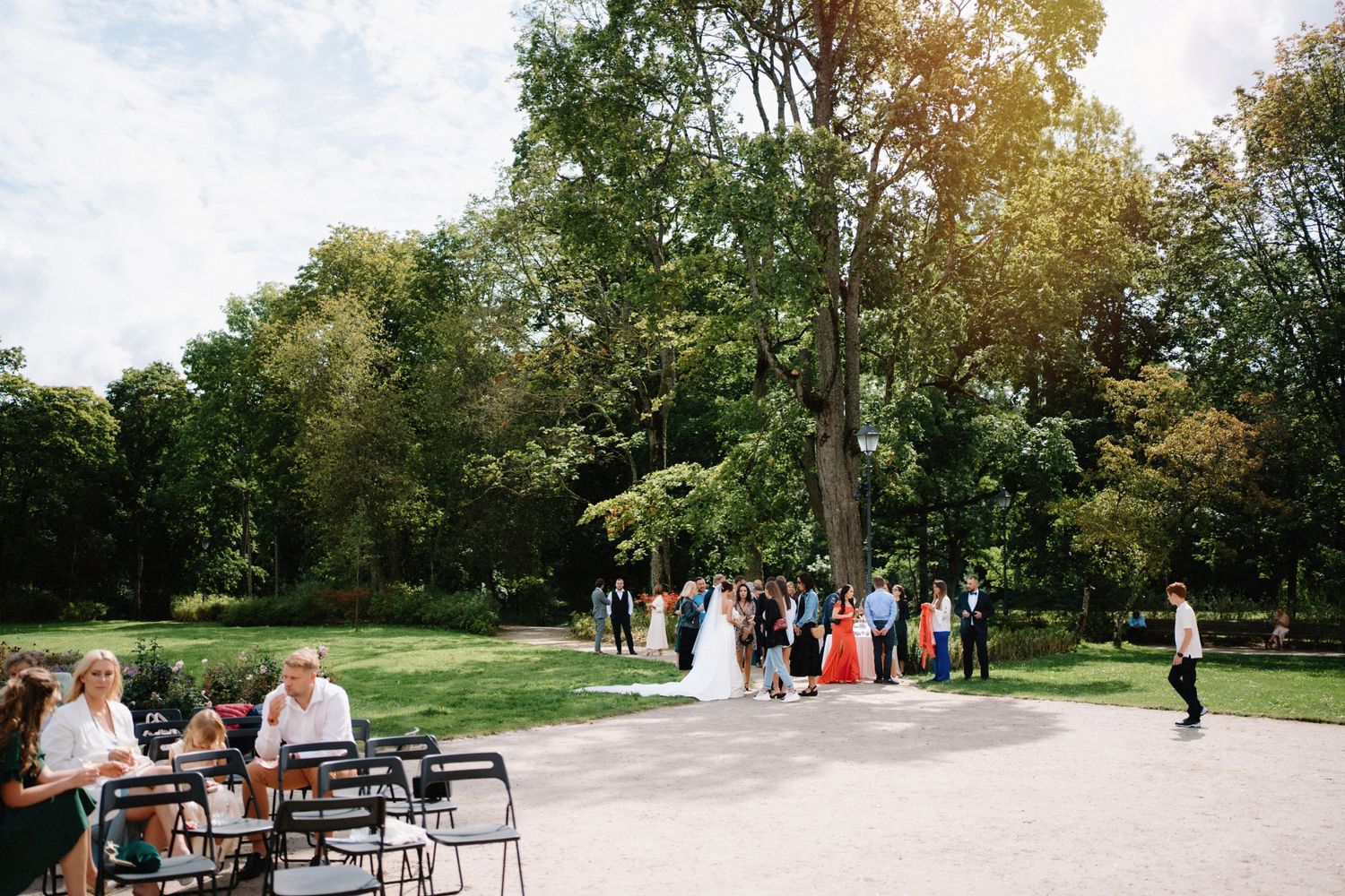 Panoramic view of wedding guests gathered on a sunny lawn with autumn trees.