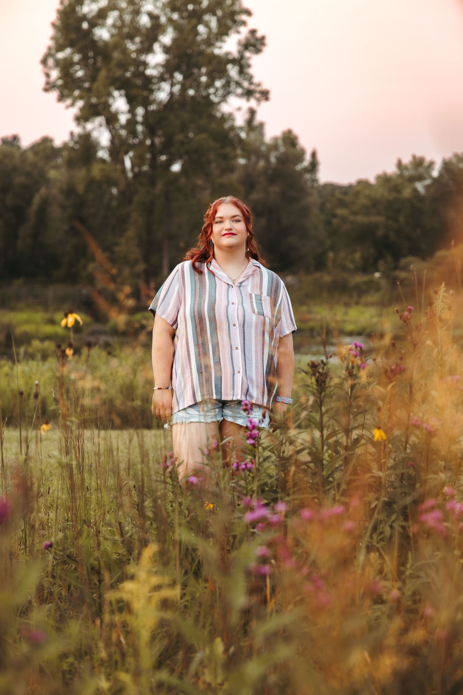 Someone in a striped button-down shirt stands in a summer meadow with wildflowers during golden hour. Metro Detroit Senior Photographer