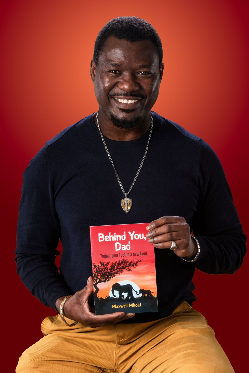 Man smiling and holding a book titled Behind You Dad by Maxwell Mkali against a red background.