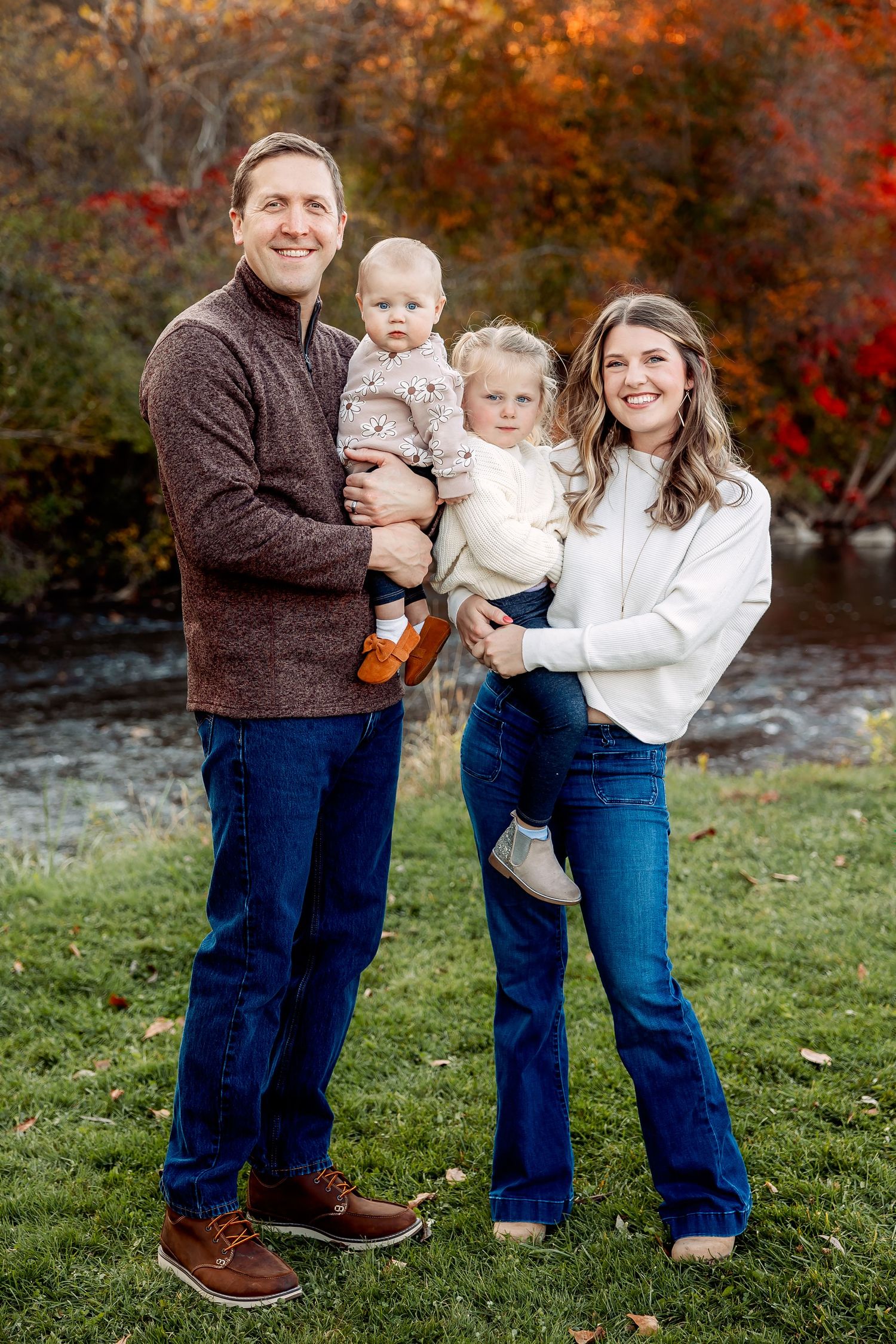 A family of four poses outdoors during autumn with colorful fall foliage in the background. Novi Northville Portrait Photographer