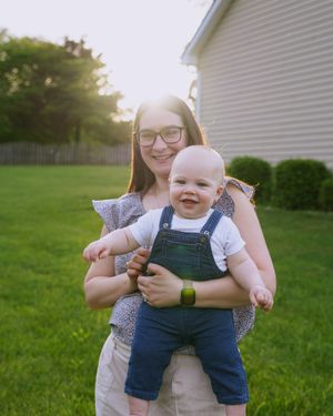 Baby in denim overalls being held in sunny backyard.