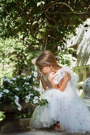 A young girl in a white tulle dress smells flowers in a garden setting.