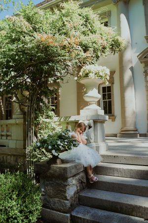 A figure in a white dress sits on stone steps of a historic building surrounded by greenery.