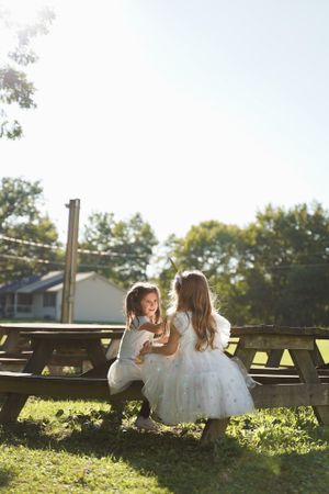 Two children in white dresses sit together on wooden benches in a sunlit garden setting.