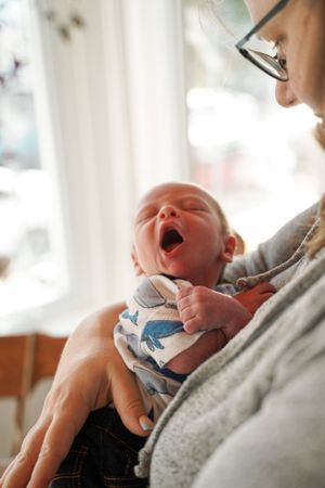 A newborn baby yawning while being held in a soft grey carrier with dinosaur print fabric.