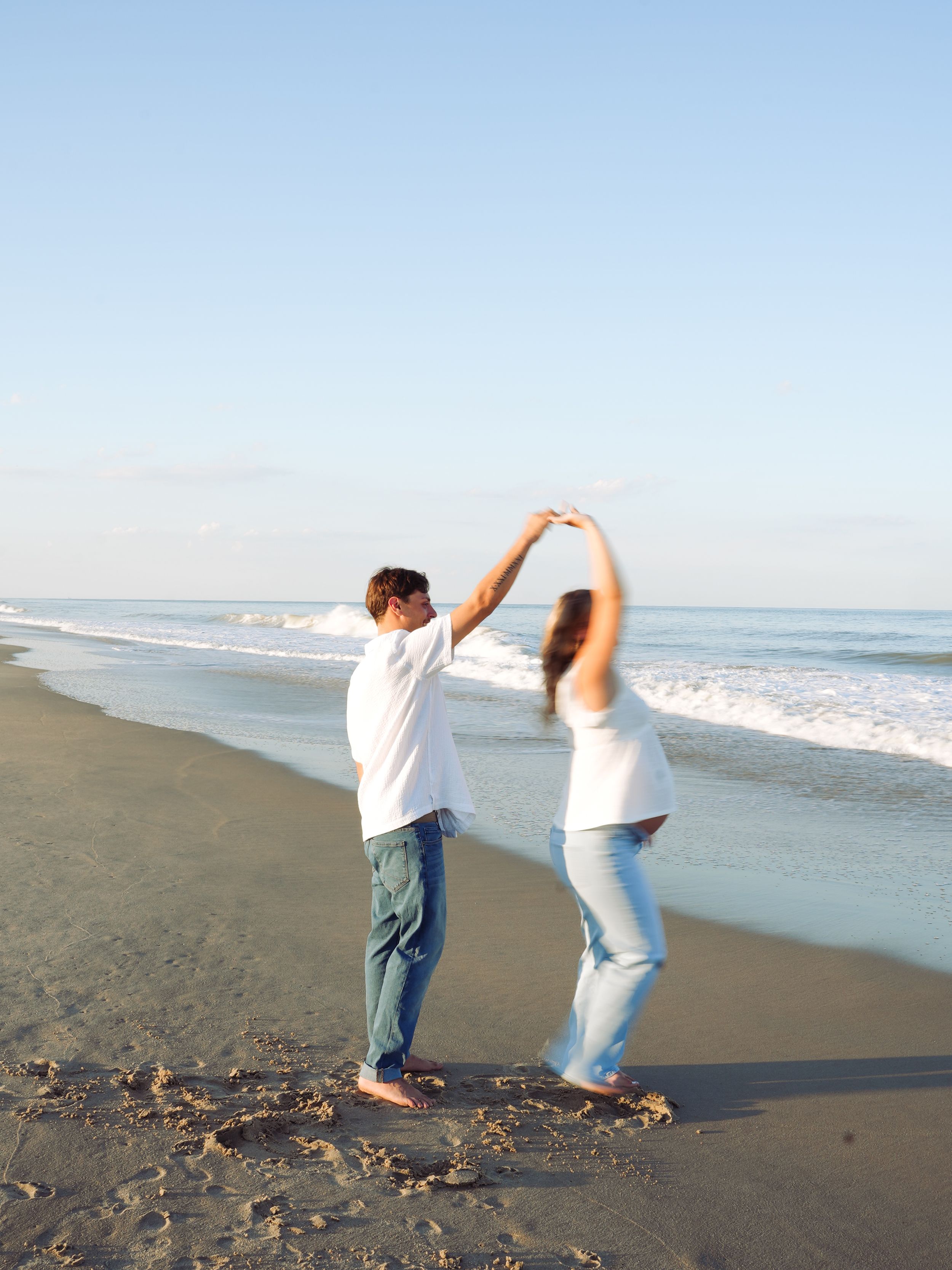 Two people playfully dance and pose together on a sandy beach with ocean waves in the background.