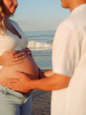 A couple in white clothing embraces on a sunny beach with ocean waves in the background.