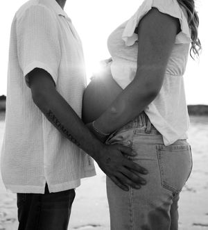 A black and white maternity photo showing a couple embracing on the beach with sunlight behind them.
