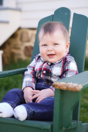 Baby wearing plaid shirt and overalls sits smiling in green Adirondack chair.