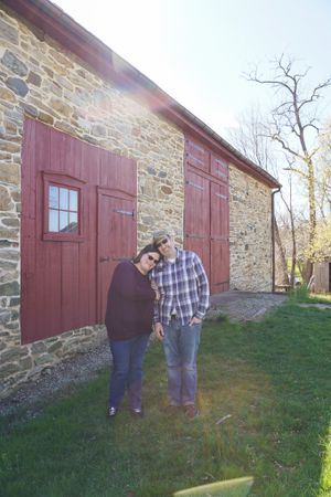 Couple standing together against rustic stone barn with red wooden doors.