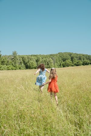 Two people in flowy dresses running through a tall grass meadow on a bright summer day.