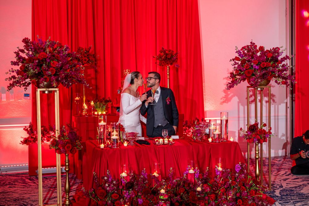 Wedding couple stands together against dramatic red backdrop with floral arrangements on tall pedestals.