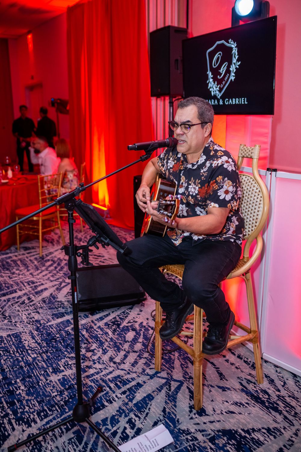Solo musician performs on acoustic guitar in floral shirt against red-lit backdrop with branded signage.