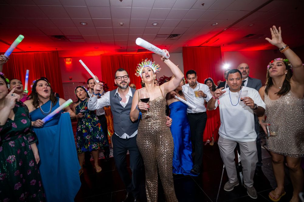 Wedding party dances enthusiastically under red lighting with glow sticks and party accessories.